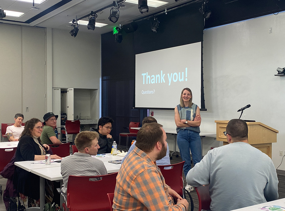 A woman stands and addresses a group of people in a classroom