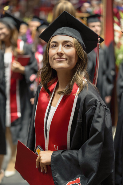 feminine student in commencement robes standing alone near a planter box