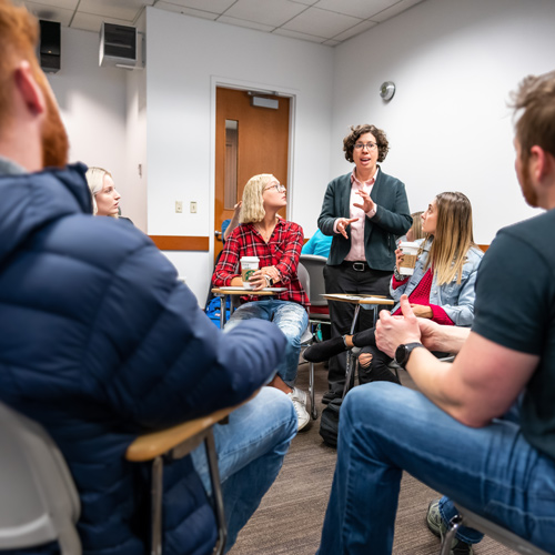 students in a classroom with their desks arranged in a circle looking at professor